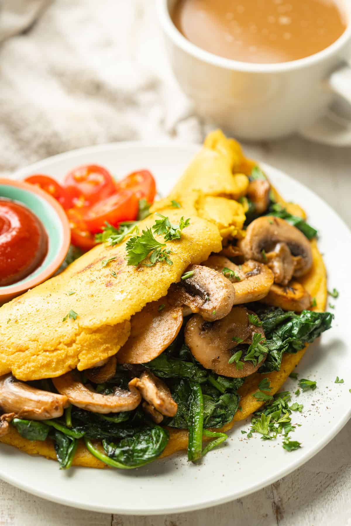 Vegan omelette stuffed with mushrooms and spinach on a plate with a cup of coffee in the background.