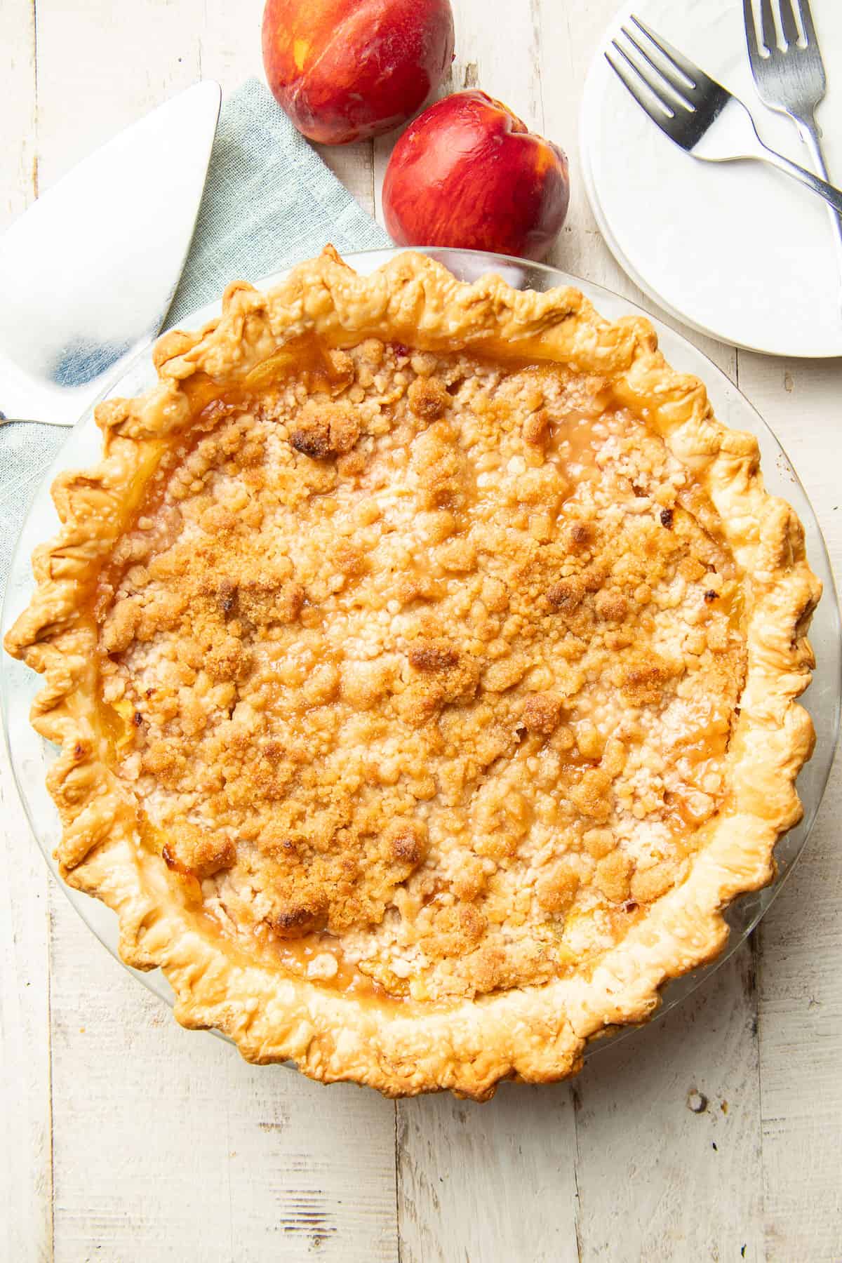 Whole Vegan Peach Pie on a white wooden surface with peaches, dishes, forks and pie server.