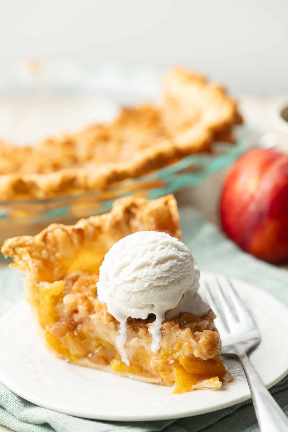 Slice of Vegan Peach Pie with vanilla ice cream, peaches and pie plate in the background.