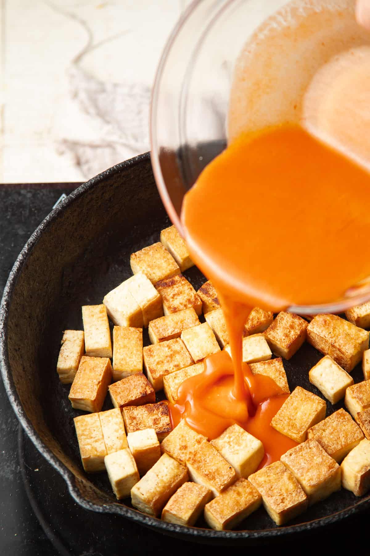 Buffalo sauce being poured over tofu cooking in a skillet.