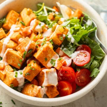 Close up of a bowl of greens and cherry tomatoes topped with Buffalo Tofu Salad and vegan ranch dressing.