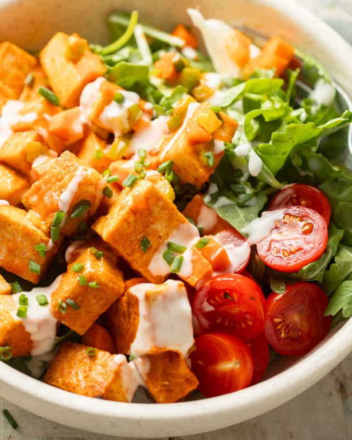 Close up of a bowl of greens and cherry tomatoes topped with Buffalo Tofu Salad and vegan ranch dressing.