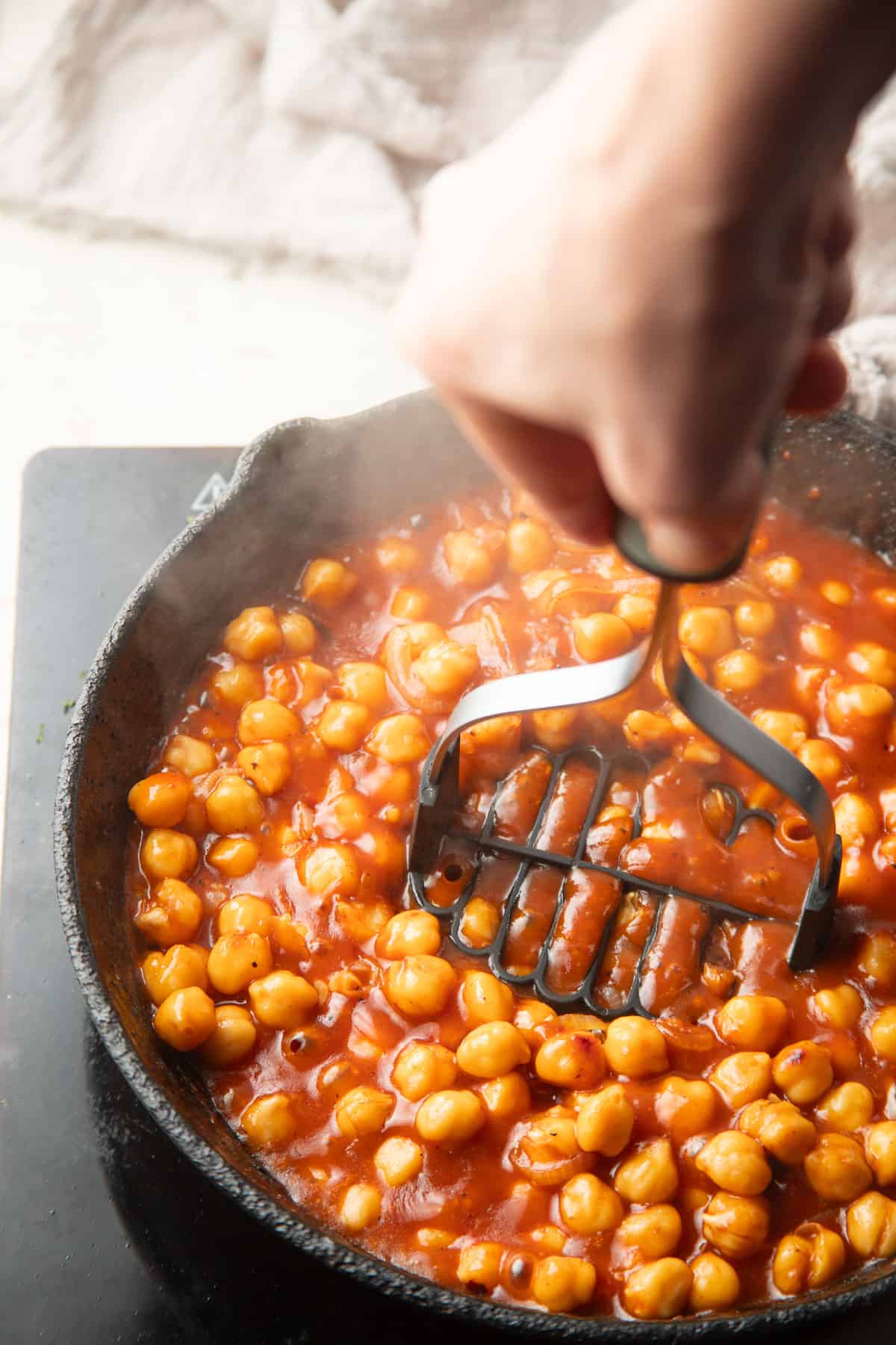 Hand mashing chickpeas in barbecue sauce in a skillet.