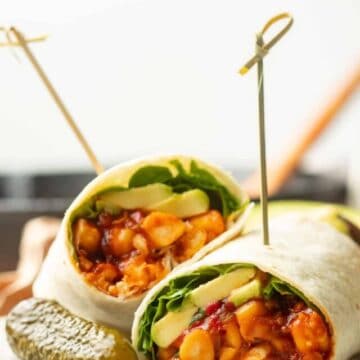 Two halves of a Barbecue Chickpea Wrap on a plate with skewers in them and a pickle on the side.