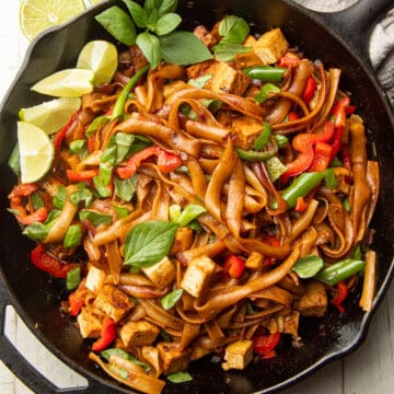 Drunken noodles in a skillet with basil and lime wedges.