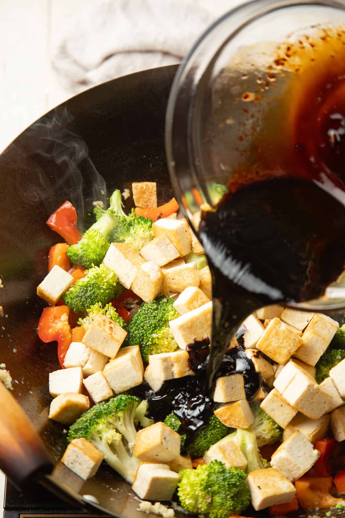Sauce being poured into a wok filled with tofu and vegetables.