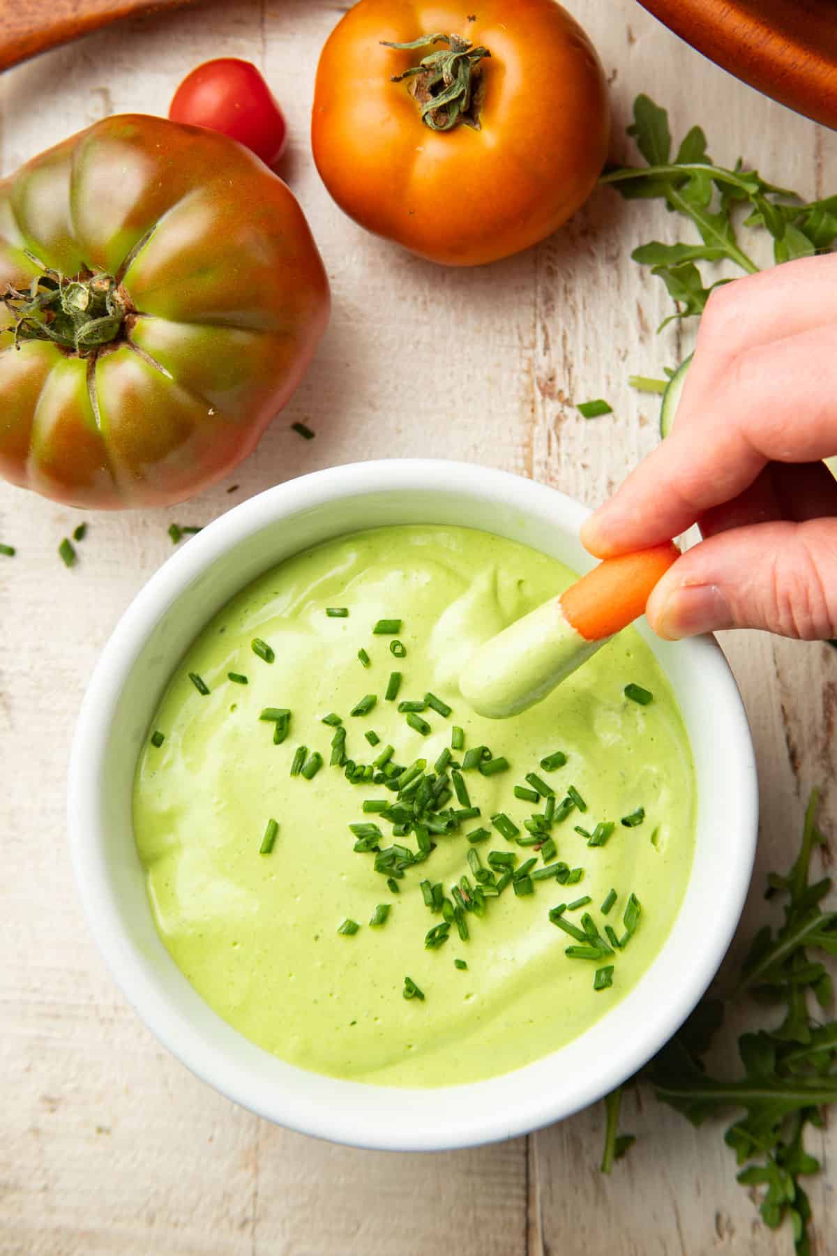 Hand dipping a baby carrot into a bowl of Vegan Green Goddess Dressing sitting on a white wooden surface with fresh vegetables.