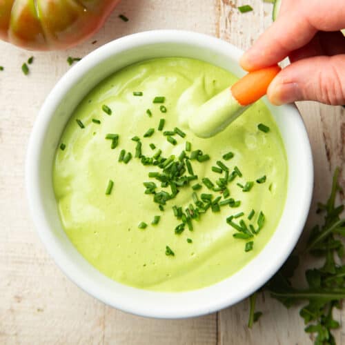 Hand dipping a carrot stick into a bowl of Vegan Green Goddess Dressing topped with chives.