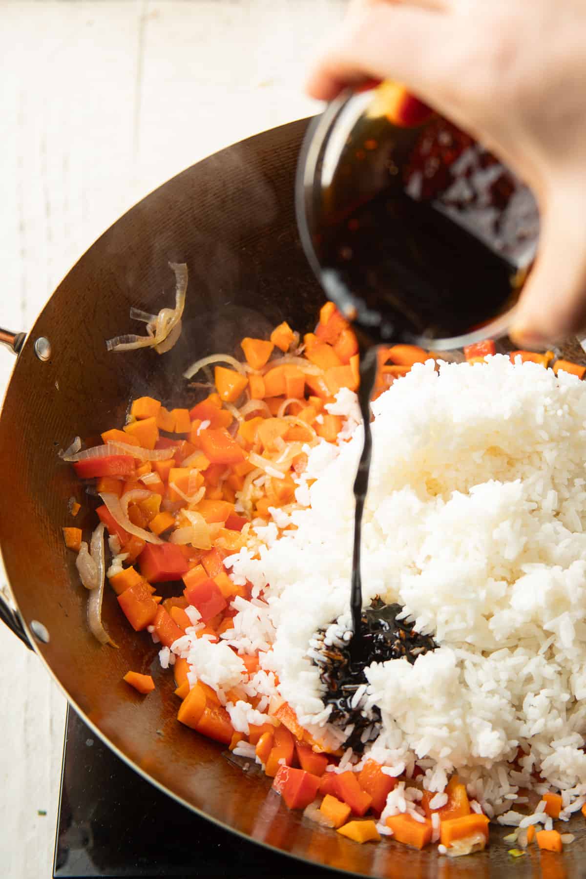 Hand pouring sauce into a wok filled with stir-fried peppers and rice.