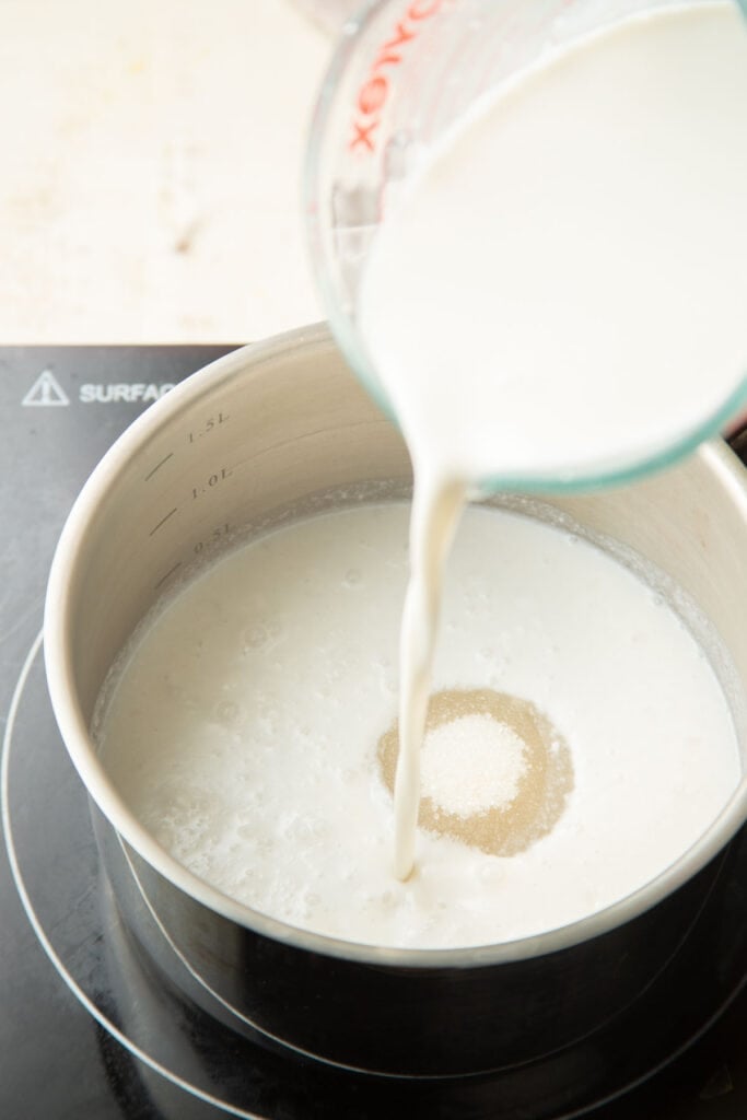 Measuring cup pouring a cornstarch slurry into a pot of coconut milk and sugar.