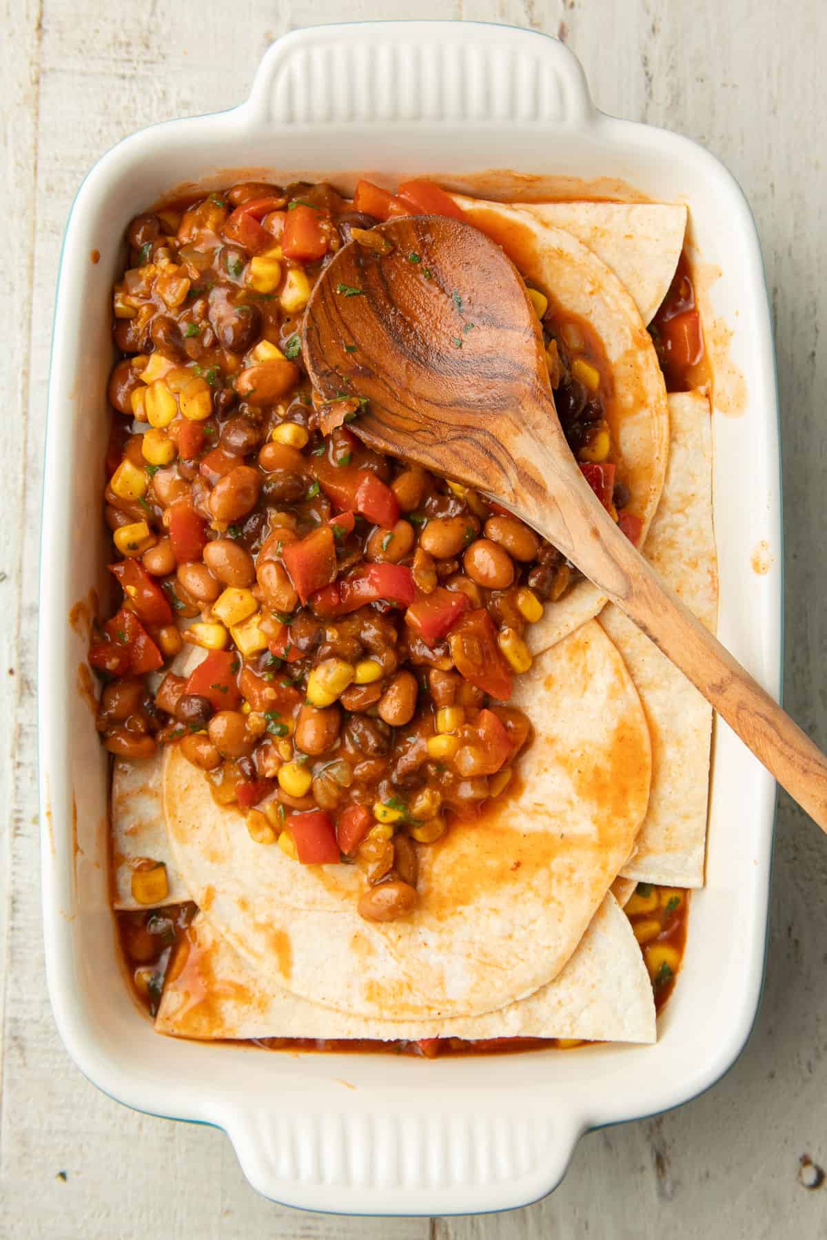 Wooden spoon spreading a layer of enchilada casserole filling over tortillas in a baking dish.