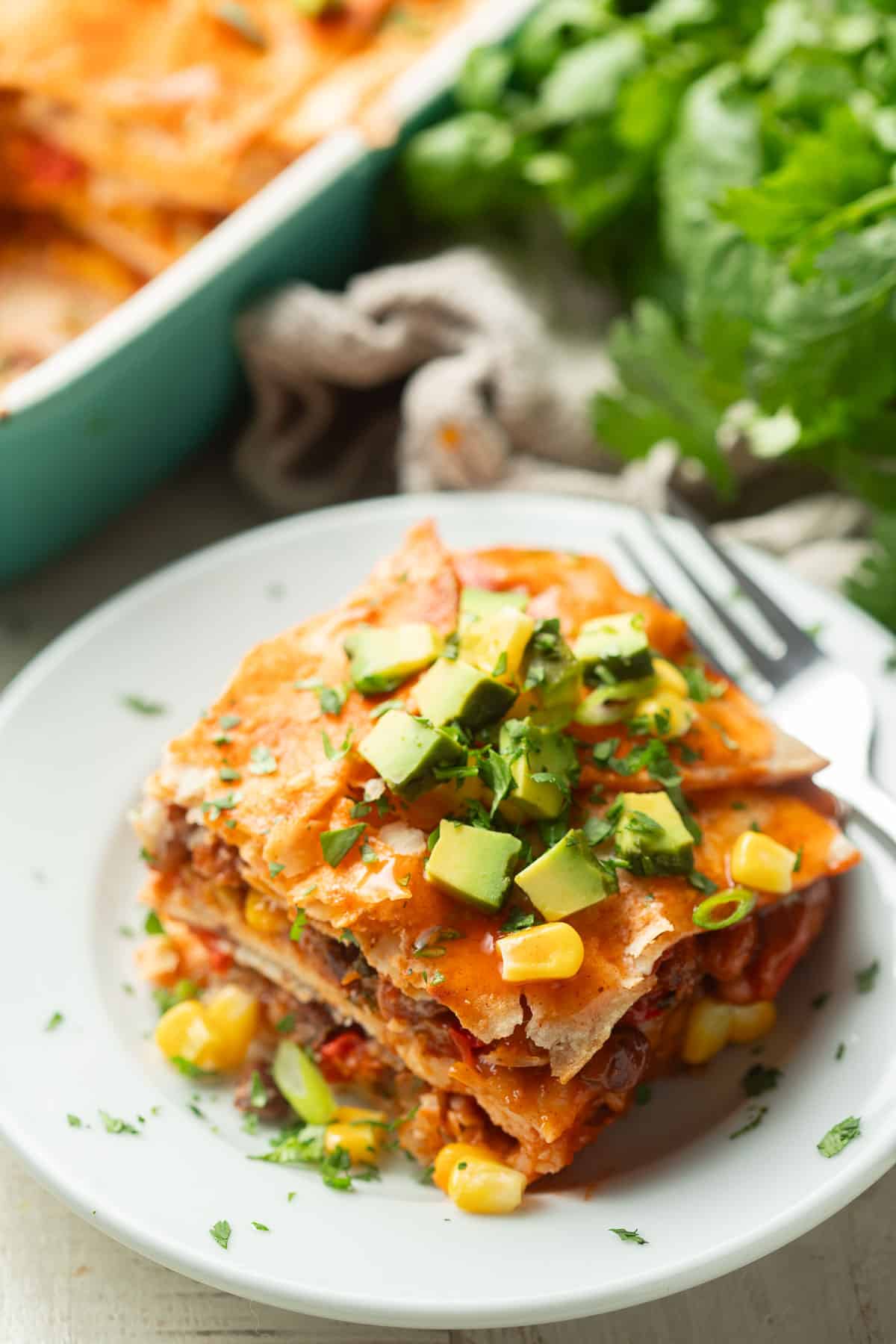 Slice of Vegan Enchilada Casserole on a dish with fork, baking dish and bunch of fresh cilantro in the background.
