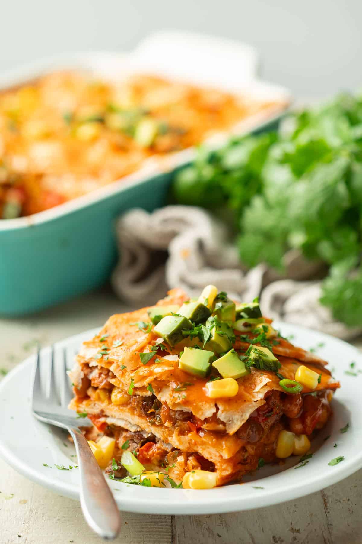 Slice of Vegan Enchilada Casserole on a plate with baking dish and bunch of cilantro in the background.