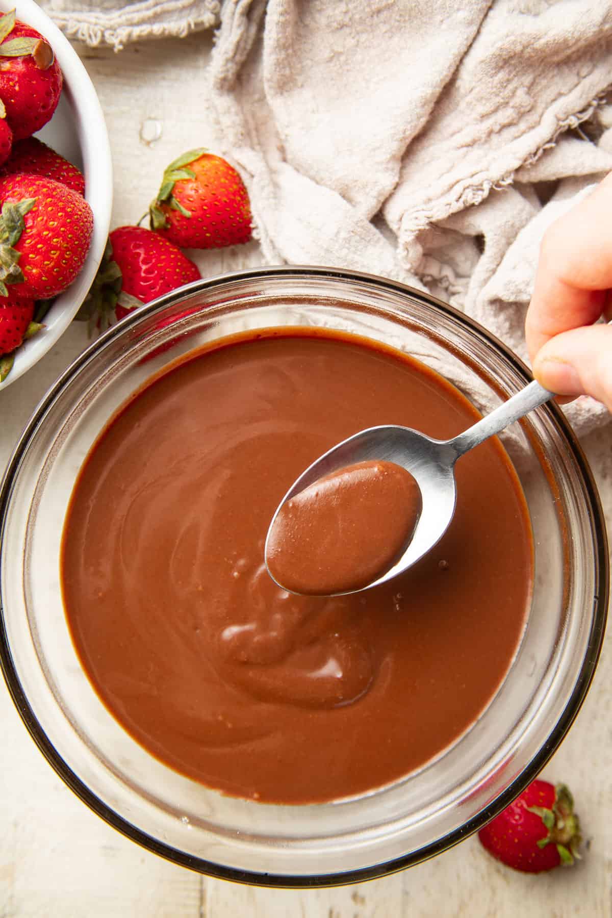 Spoon held over a bowl of Vegan Ganache on a table with fresh strawberries.