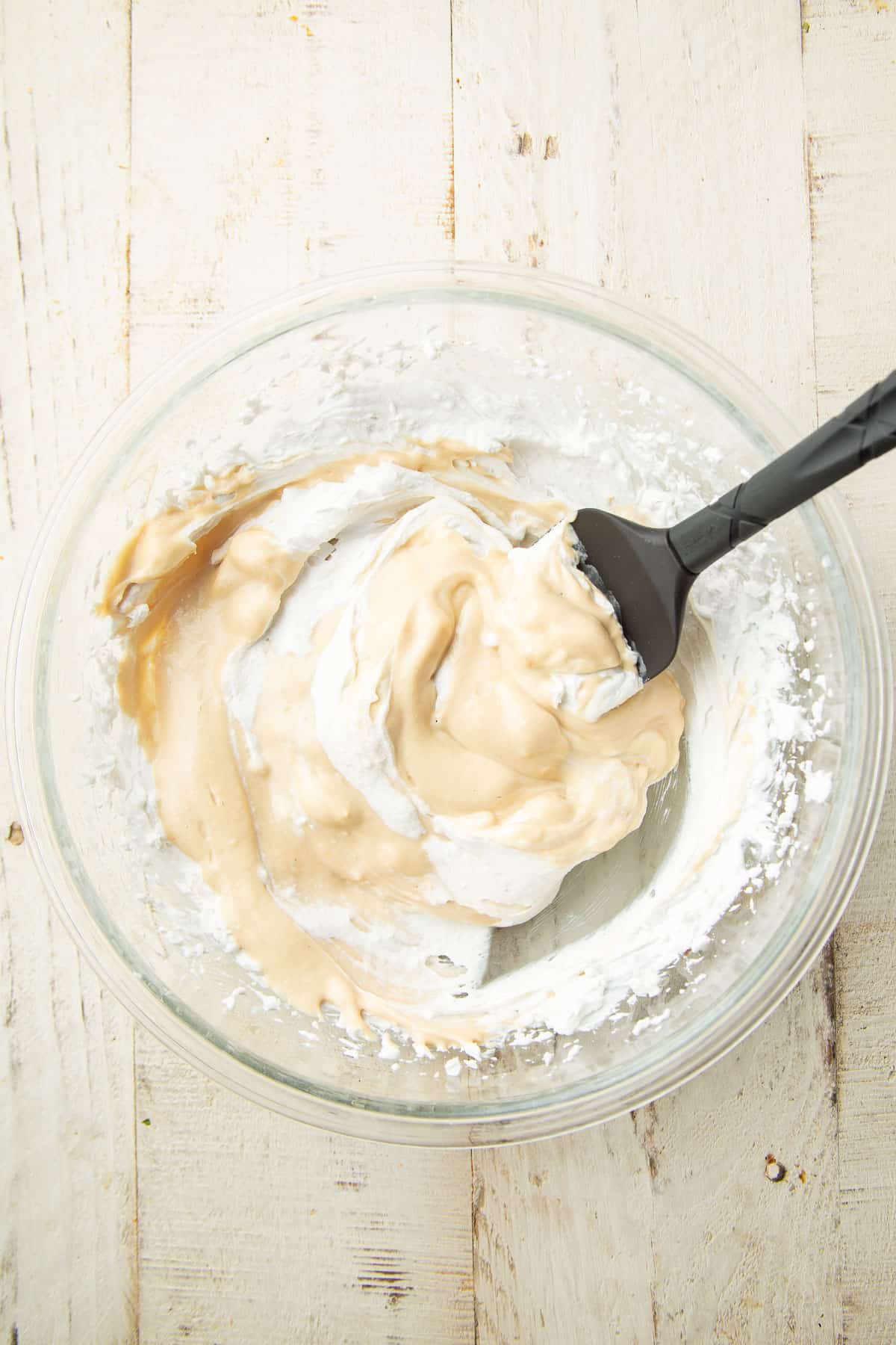 Bowl containing partially combined cashew cheese and whipped coconut cream with a spatula.
