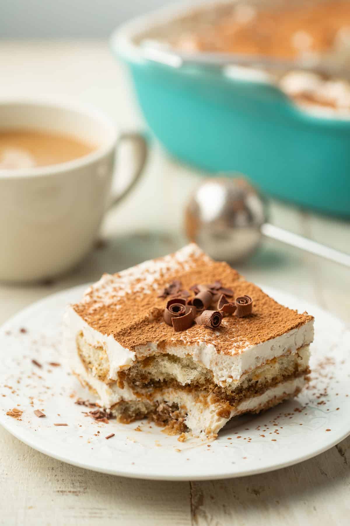 Slice of Vegan Tiramisu on a plate with coffee cup and blue baking dish in the background.