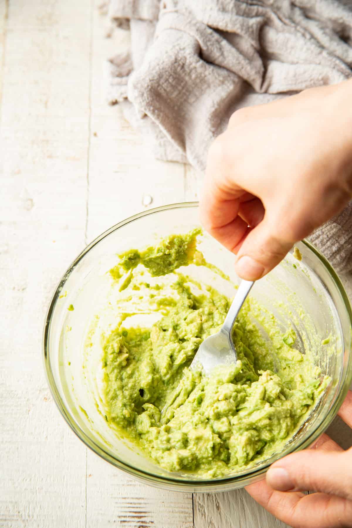 Hand stirring avocado mash together in a glass bowl.