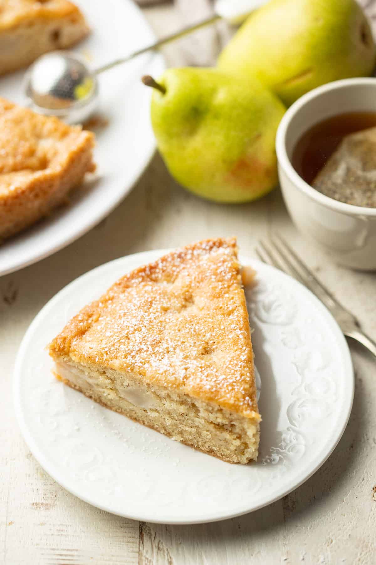 Slice of Vegan Pear Cake on a dish with tea cup and pears in the background.