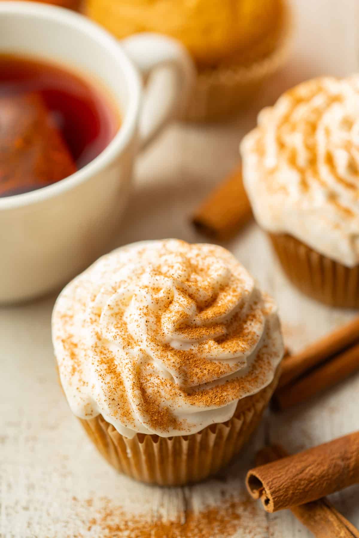Vegan Pumpkin Cupcakes on a white wooden surface with cinnamon sticks and a cup of tea.