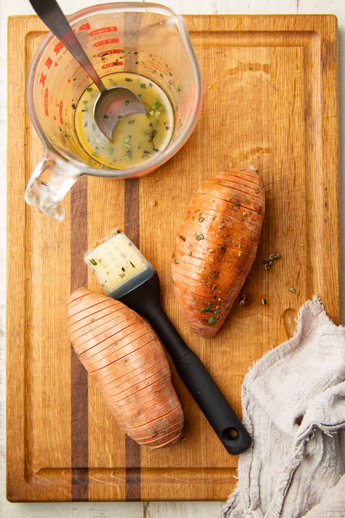 Cutting board with two uncooked Hasselback Sweet Potatoes, basting brush, and measuring cup filled with rosemary butter.
