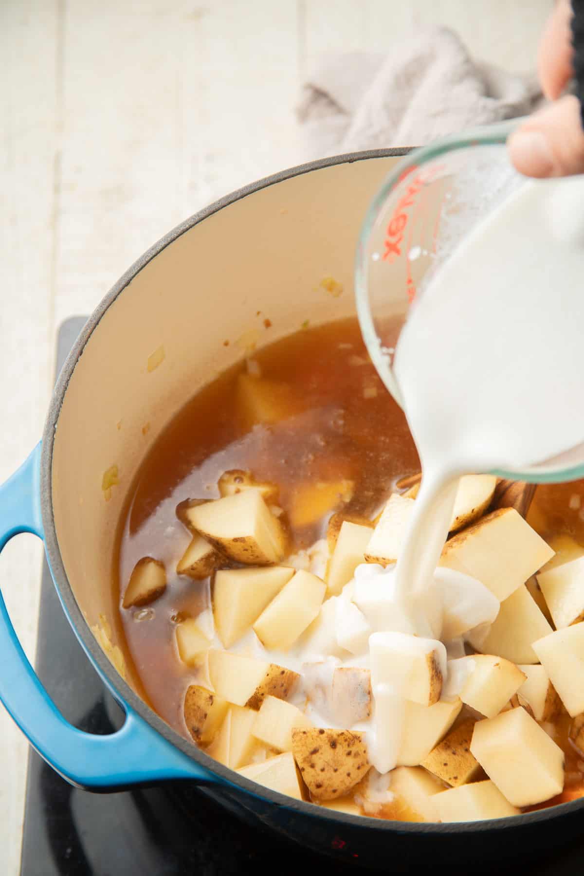 Hand pouring coconut milk into a pot of potatoes and broth.