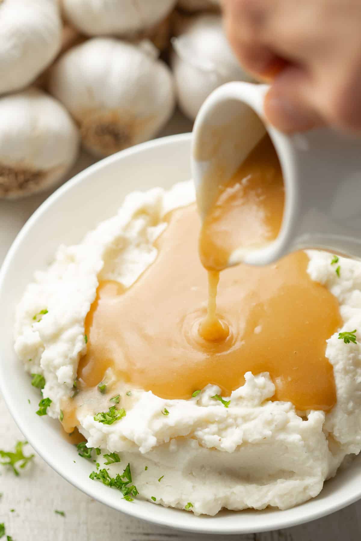 Hand pouring gravy over a bowl of mashed potatoes.