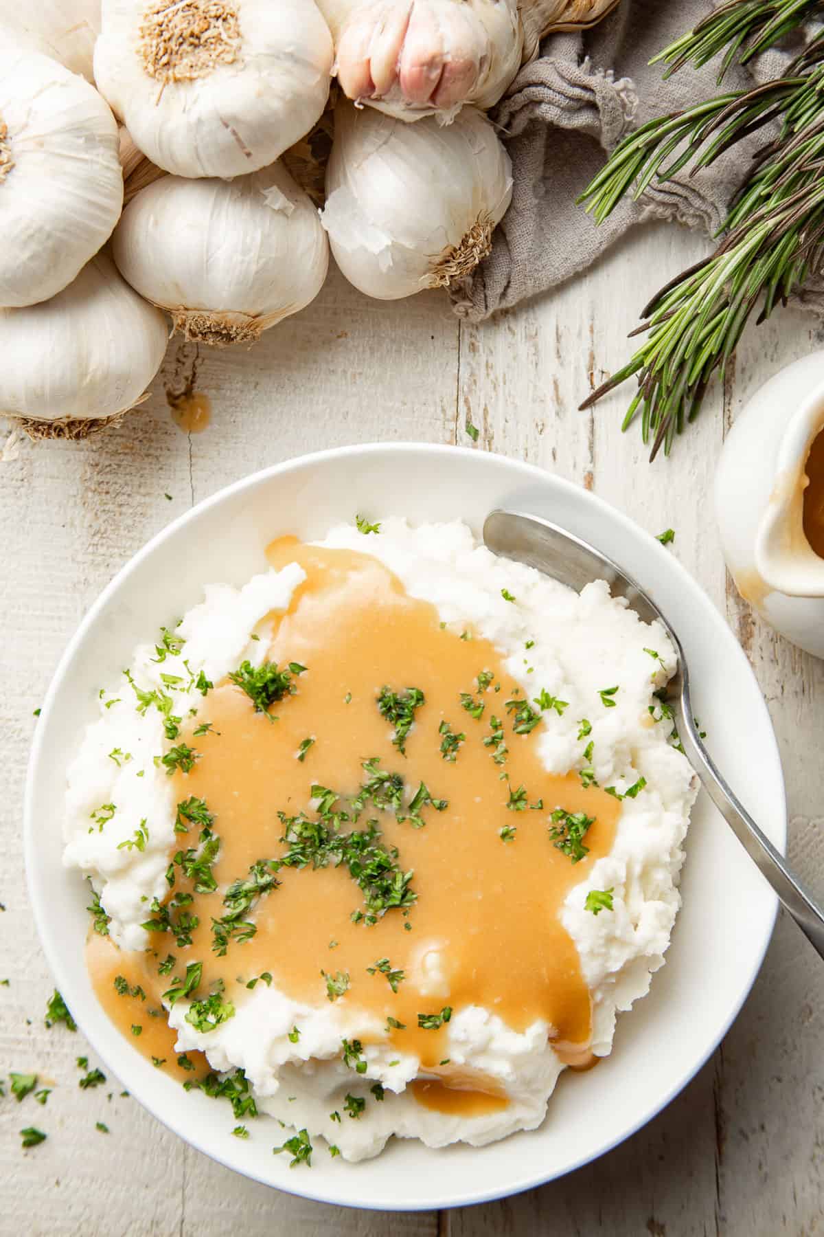 White wooden surface set with fresh garlic and rosemary sprigs, and bowl of mashed potatoes and Roasted Garlic Gravy.
