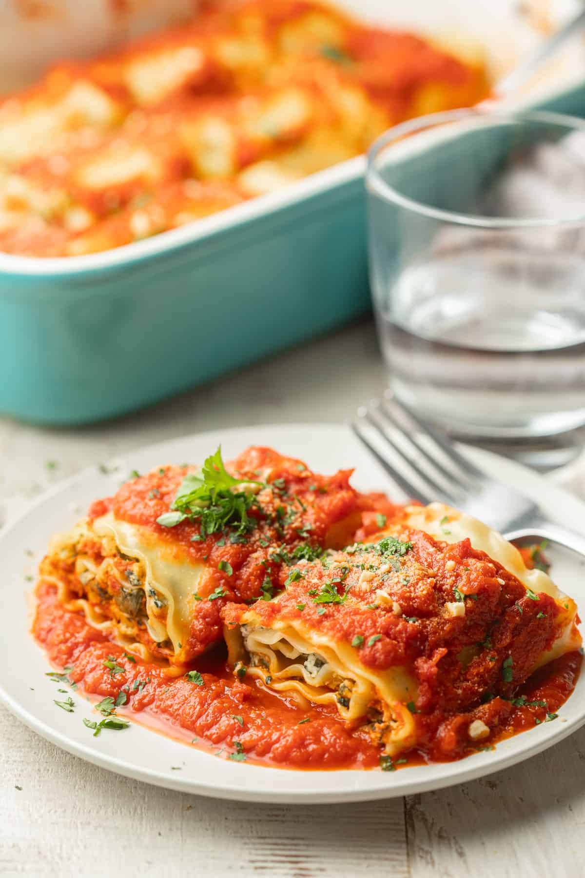 Two Vegan Lasagna Roll-Ups on a place with water glass and baking dish in the background.