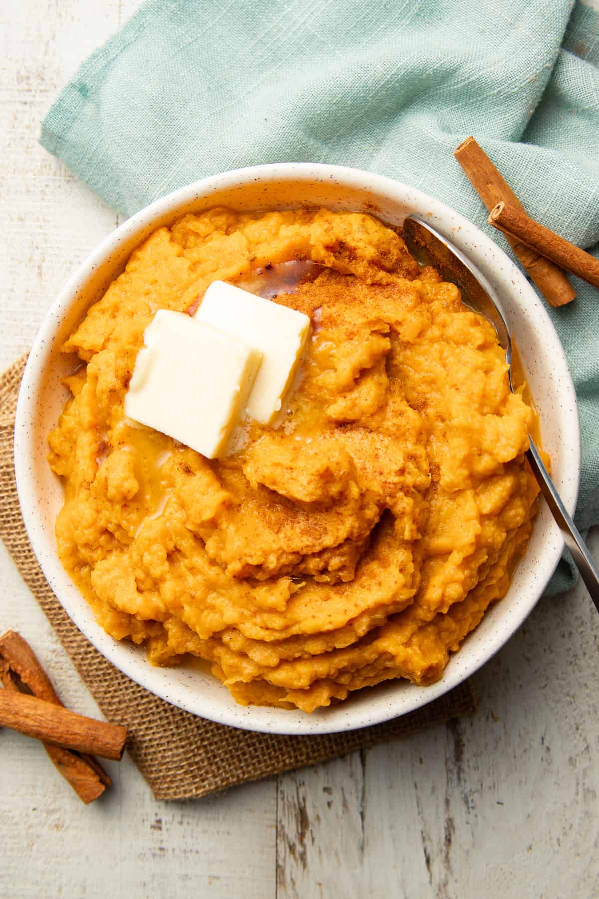 White wooden surface set with a blue napkin, cinnamon sticks and a bowl of Vegan Mashed Sweet Potatoes.