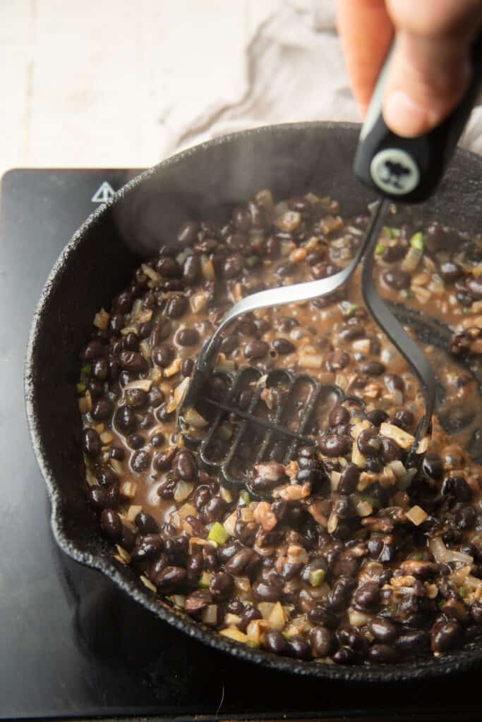Hand mashing black beans in a skillet with a potato masher.