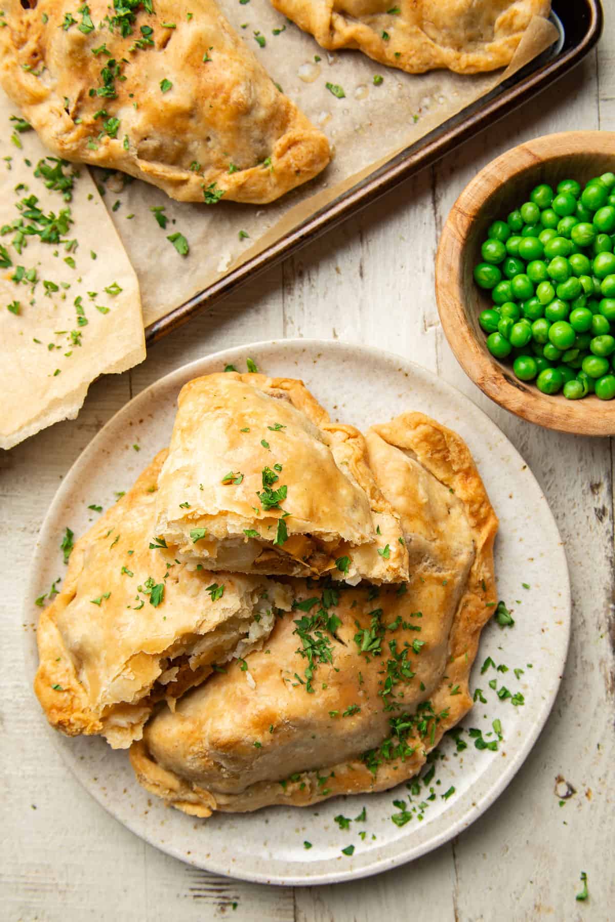 White wooden surface set with baking sheet, bowl of peas and plate of Vegan Cornish Pasties.
