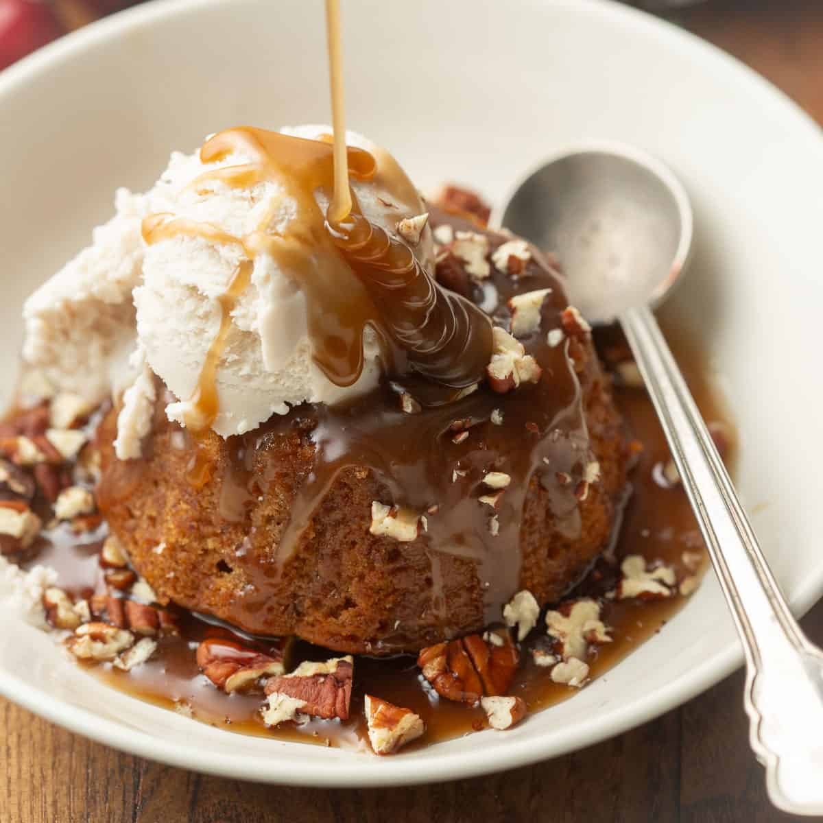 Vegan Sticky Toffee Pudding in a bowl with ice cream and nuts, toffee sauce being drizzled on top.
