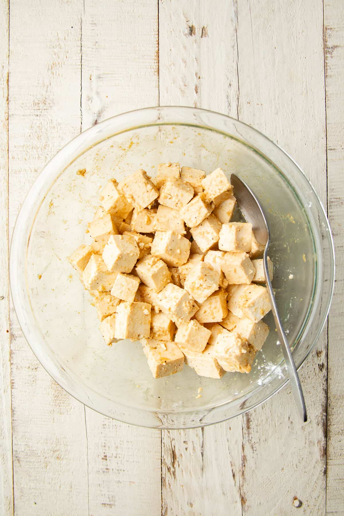 Tofu cubes coated in seasonings, oil and cornstarch, in a glass bowl with spoon.