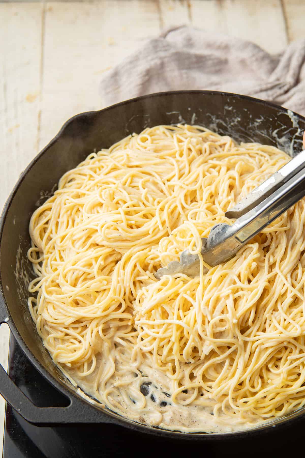 Vegan Cacio e Pepe cooking in a skillet with pair of tongs.