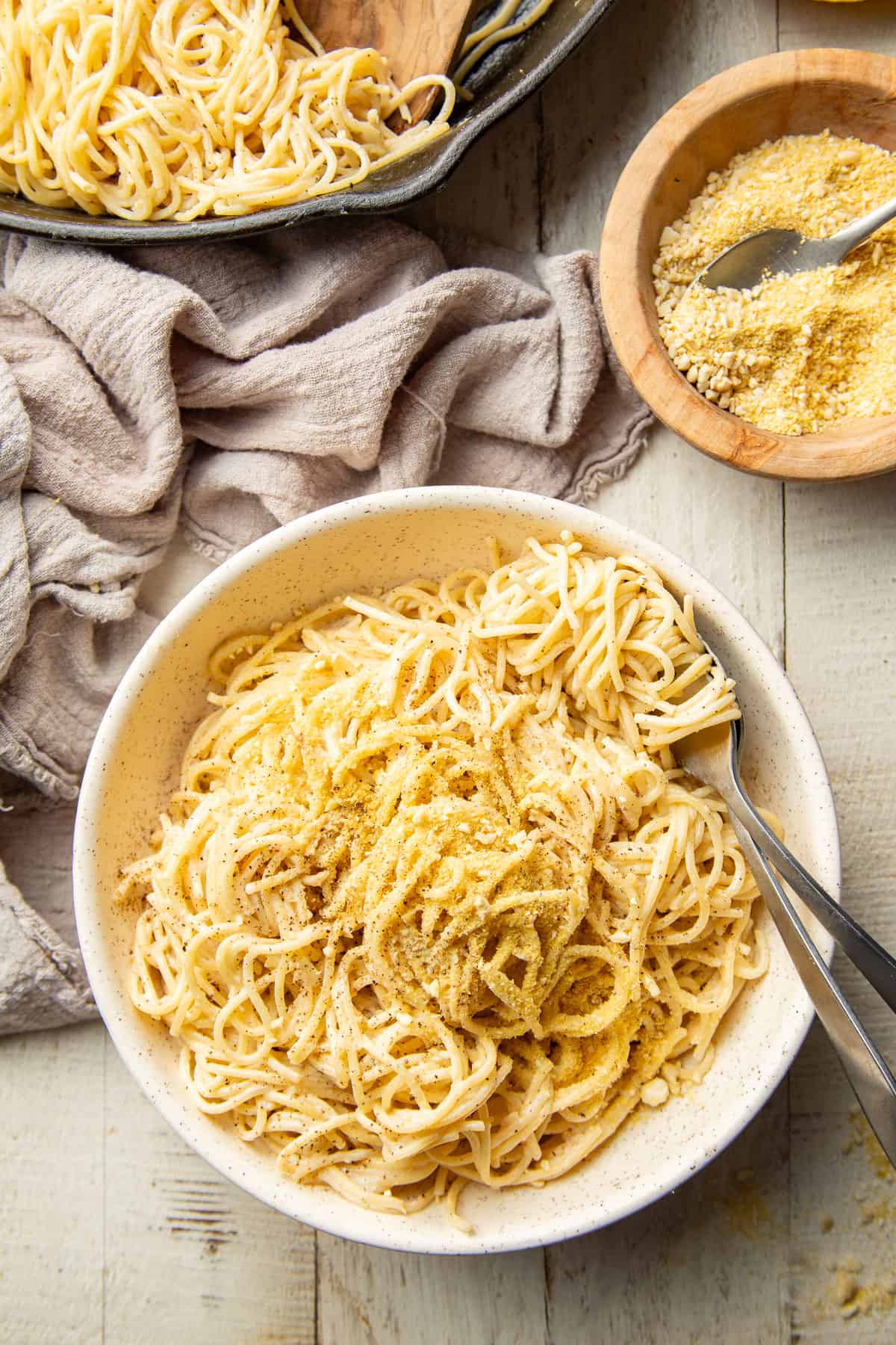 White wooden surface set with skillet, bowl of vegan Parmesan cheese and bowl of Vegan Cacio e Pepe.