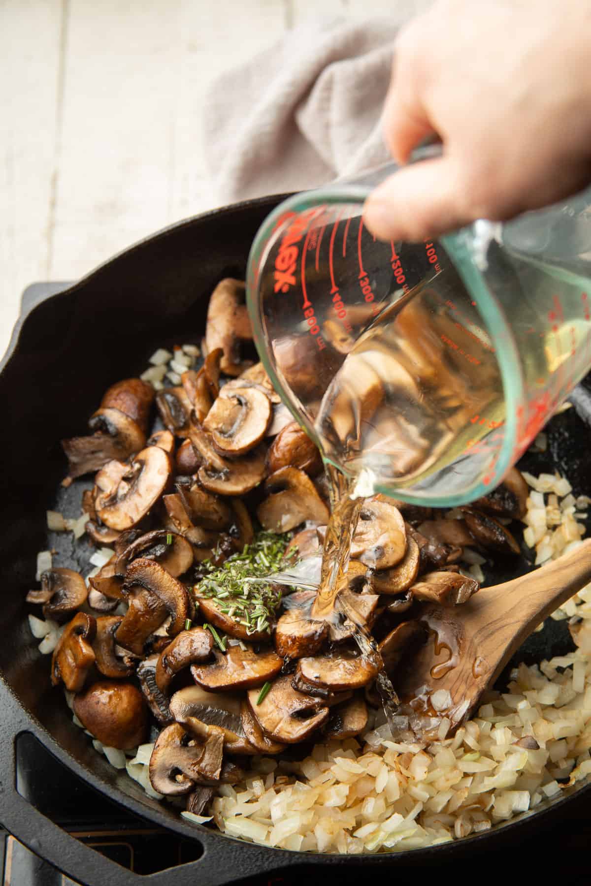 Hand pouring dry sherry into a skillet of mushrooms, onions and rosemary.