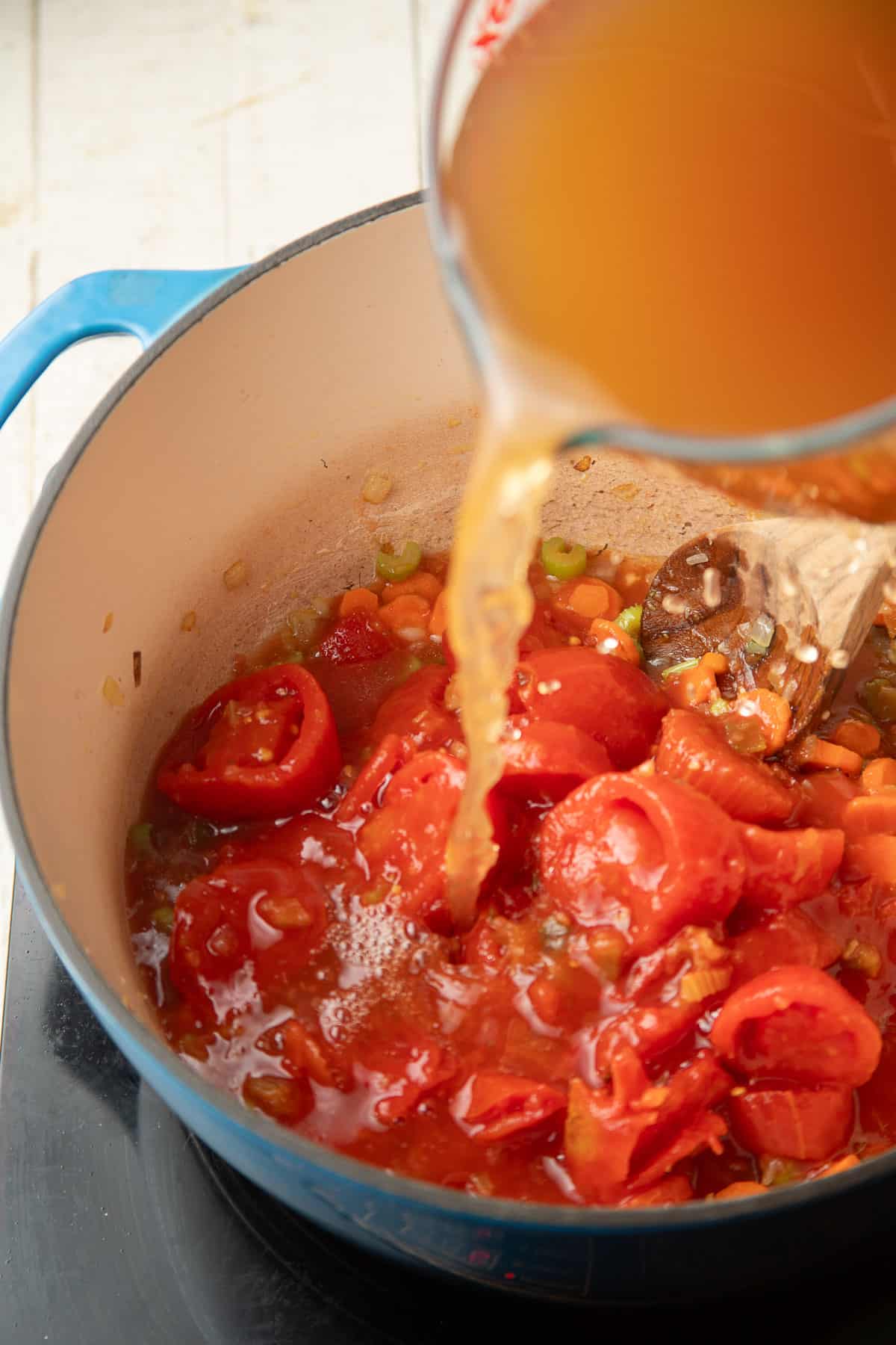 Broth being pouted into a pot containing tomatoes and vegetables.