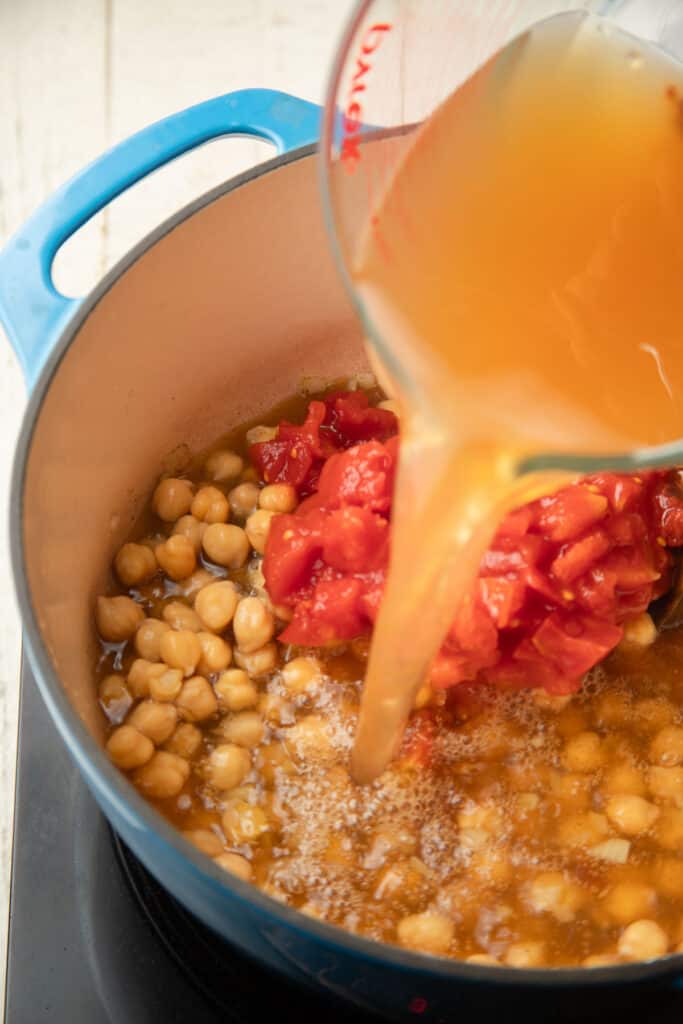 Broth being poured into a pot of diced tomatoes and chickpeas.