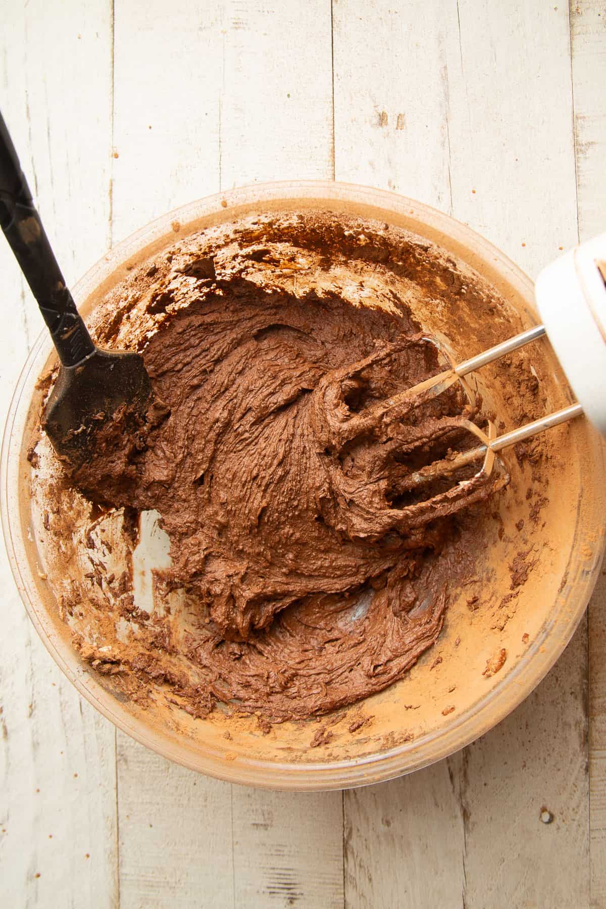 Mixture of wet ingredients and cocoa for cookie dough in a bowl with electric mixer and spatula.