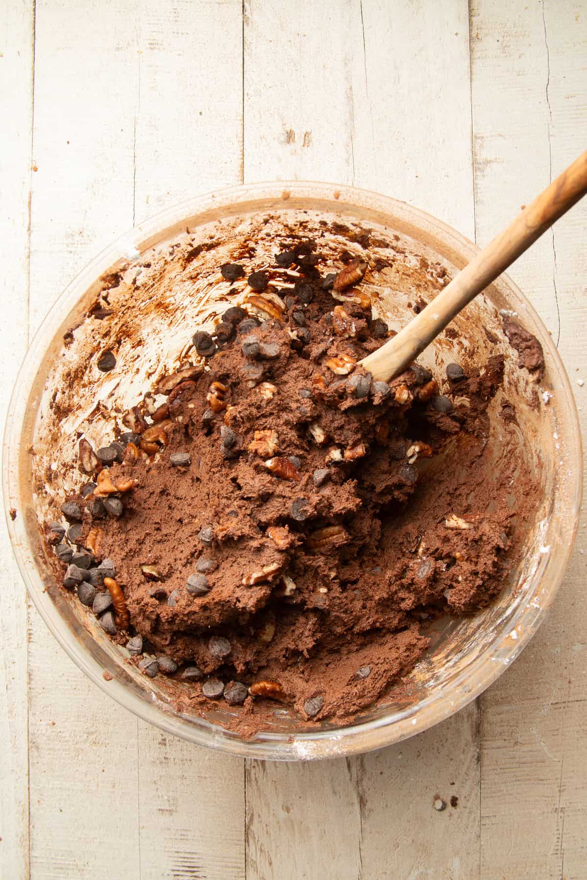 Chocolate cookie dough in bowl with chocolate chips and pecans, with a wooden spoon.