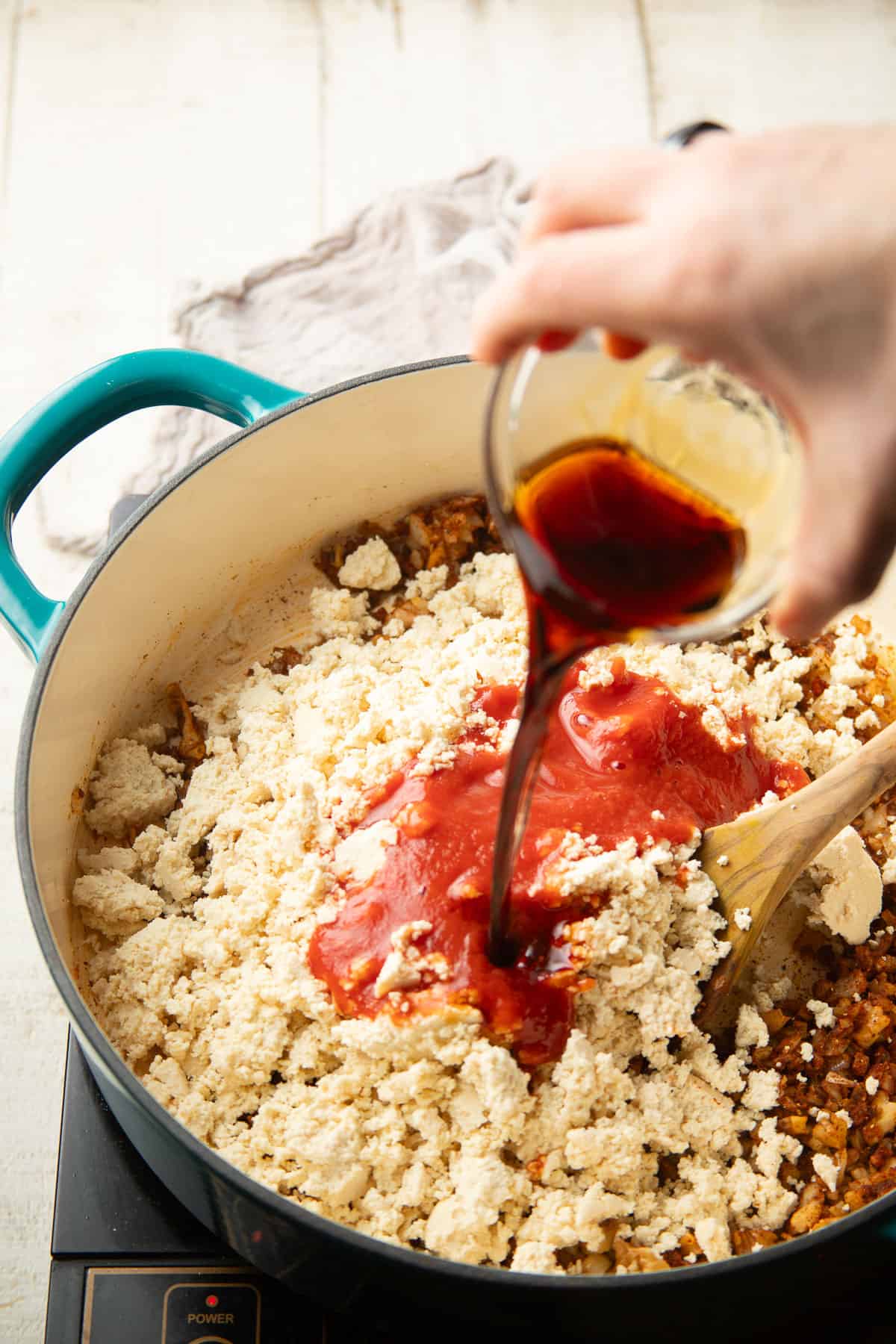 Hand pouring soy sauce into a skillet filled with crumbled tofu, walnuts, spices, and tomato sauce.