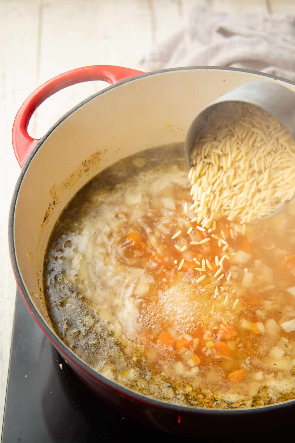 Dried orzo being poured into a pot of simmering soup.