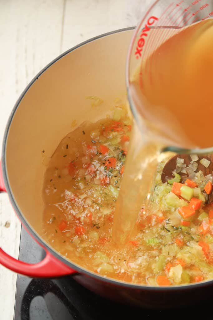 Broth being poured into a pot containing veggies and herbs.