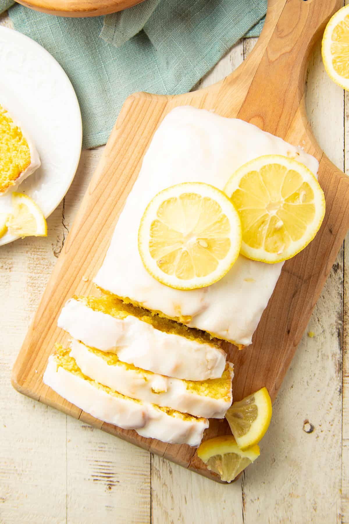 White wooden surface set with blue napkin, lemon slices, and Vegan Lemon Loaf on a cutting board.