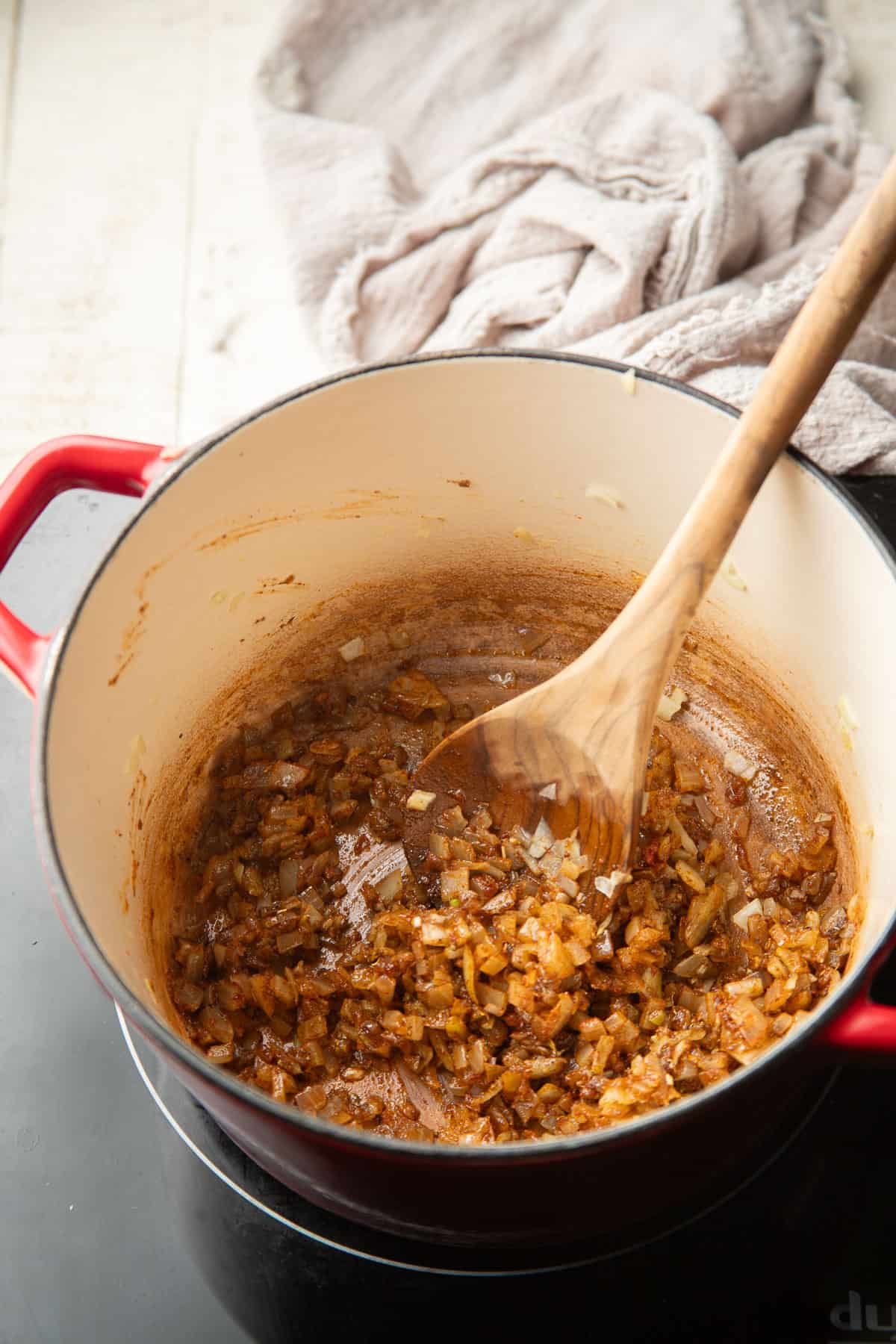 Diced onion, garlic, and spices cooking in a pot with a wooden spoon.