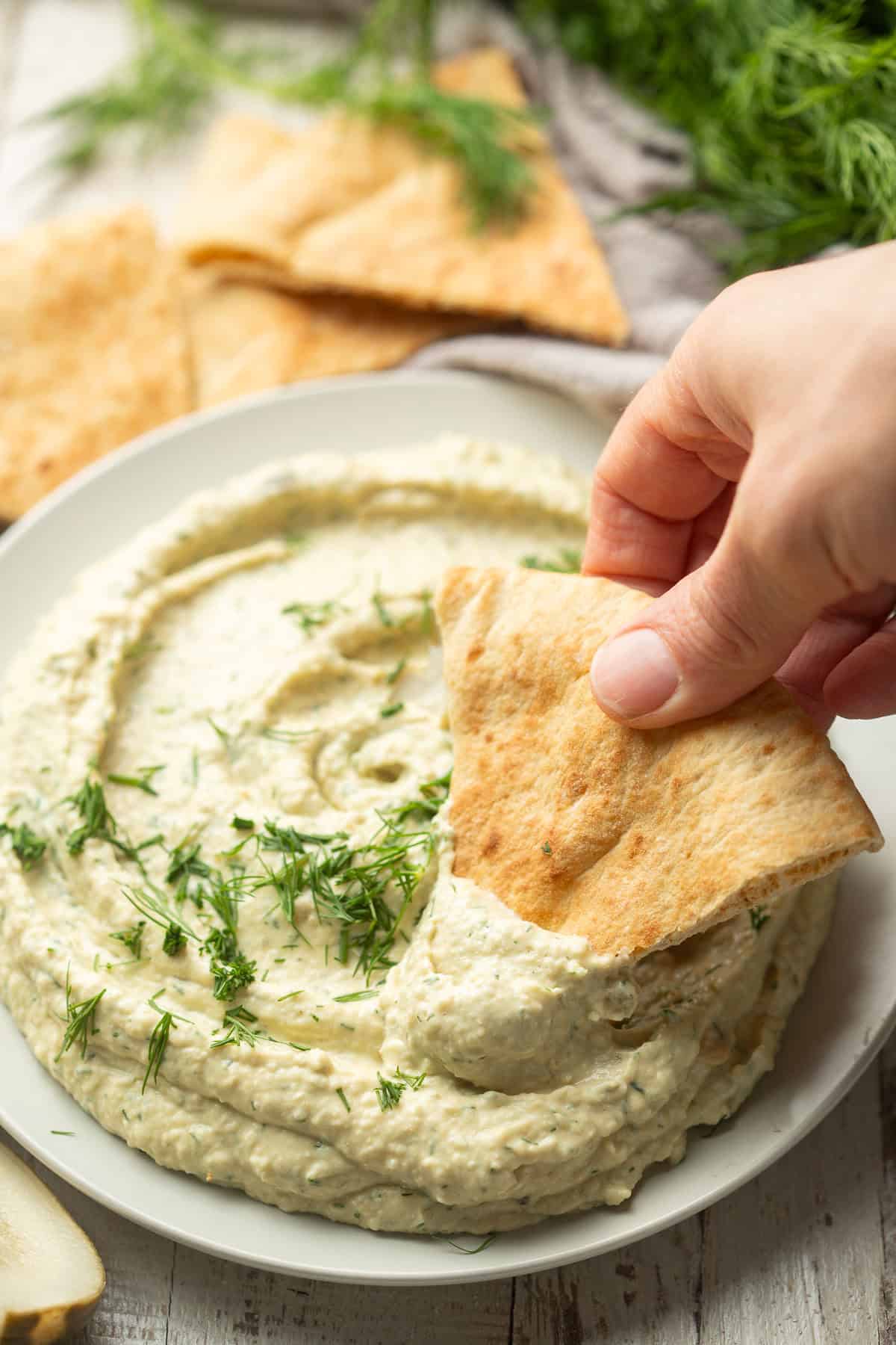 Hand dipping a pita wedge into a plate of Dill Pickle Hummus.