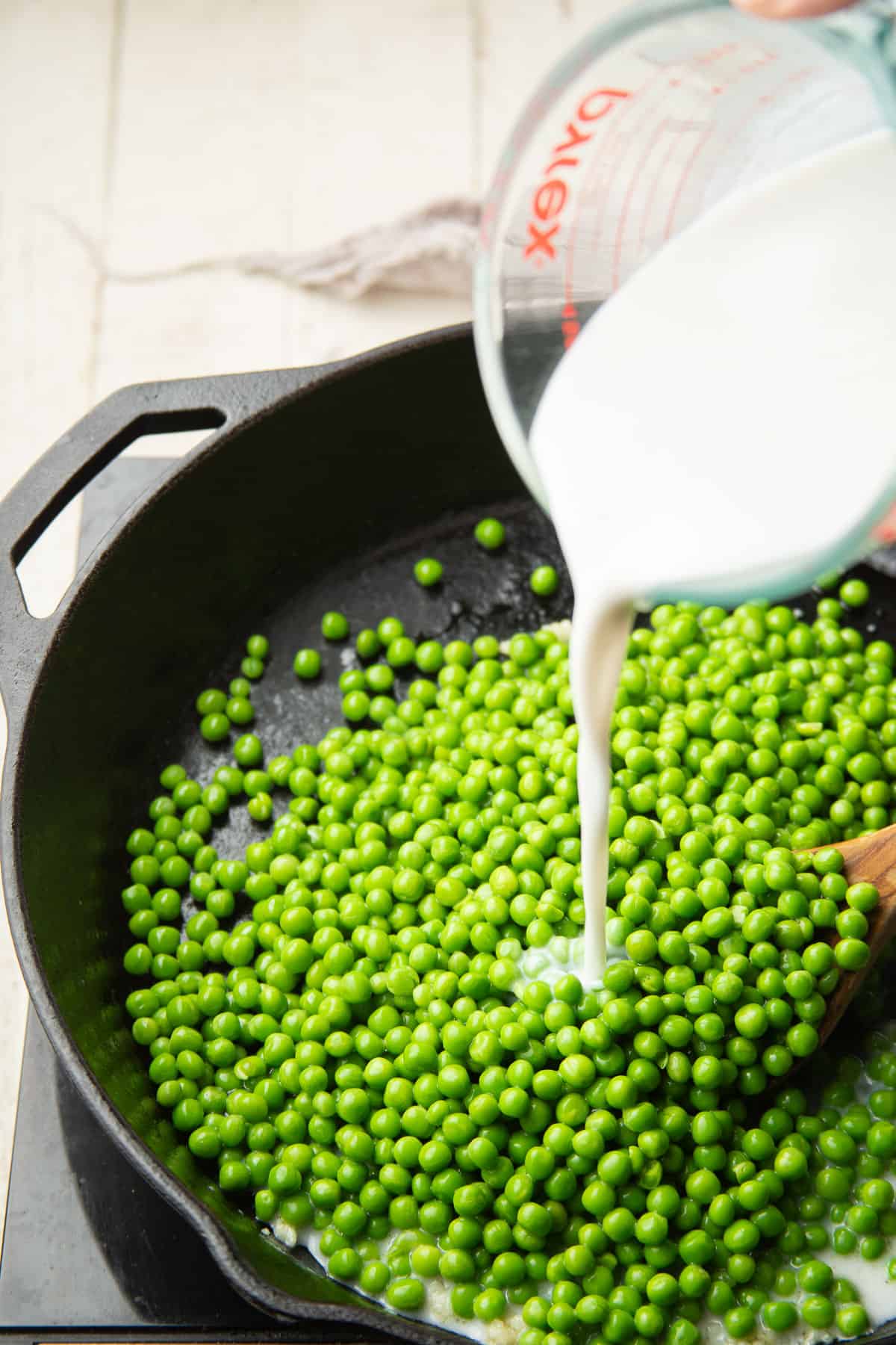 Coconut milk being poured into a skillet filled with peas.