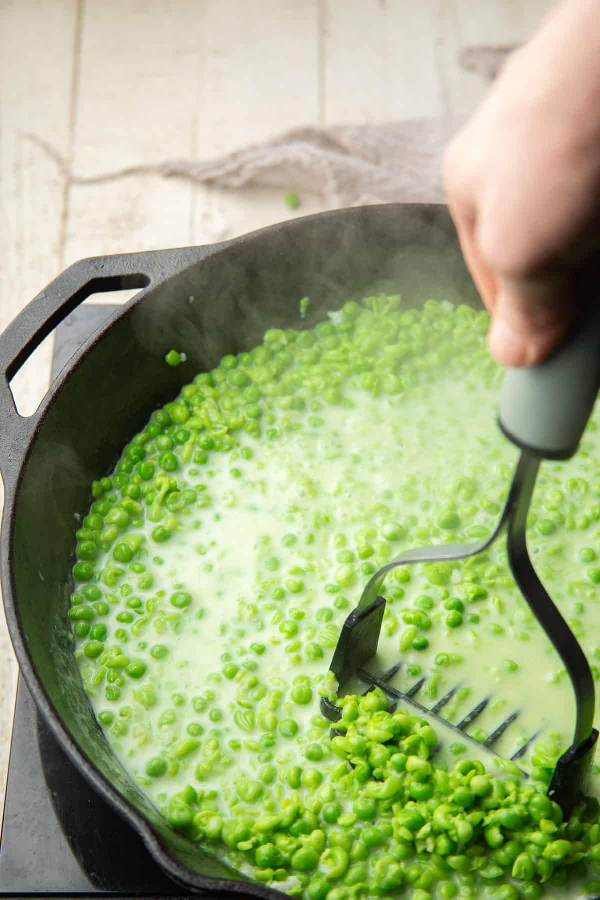 Hand with a potato masher mashing green peas in a skillet.