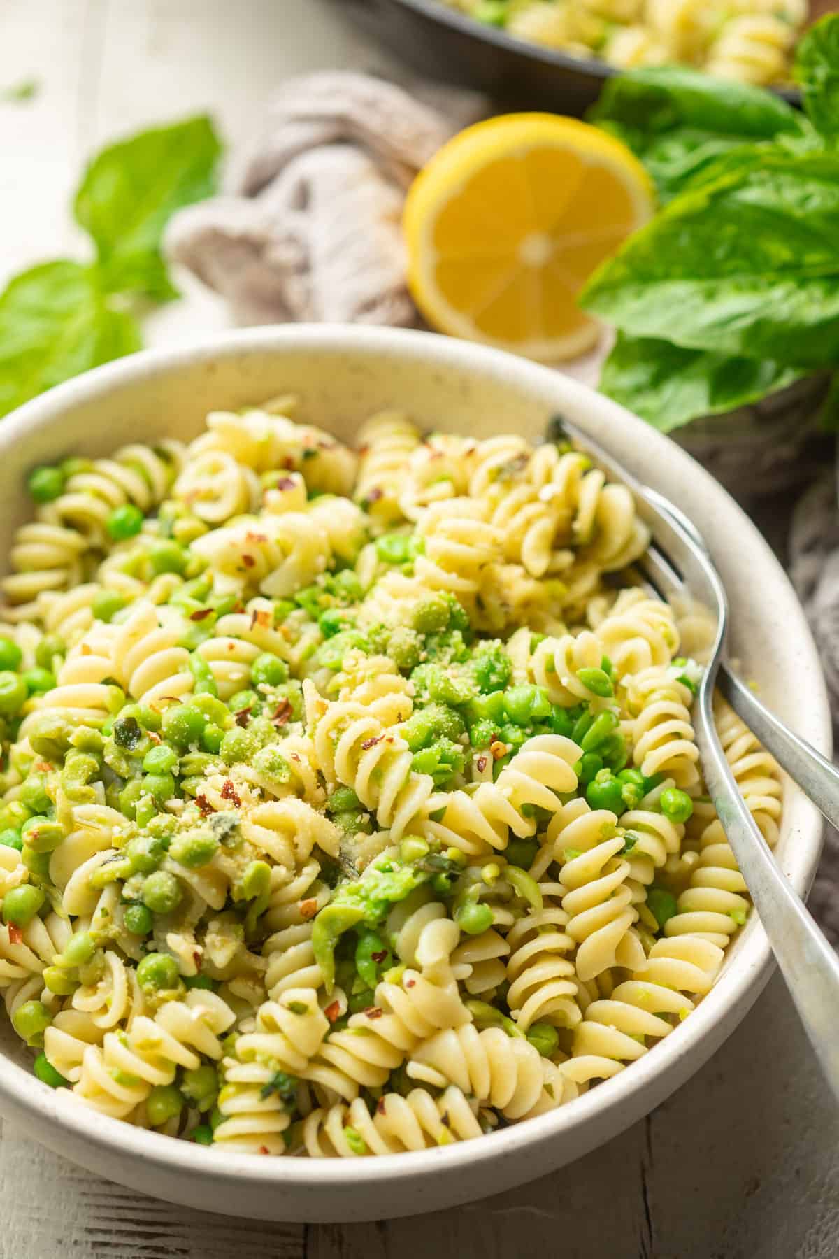 Bowl of pasta and peas with lemon half, fresh basil, and a skillet in the background.