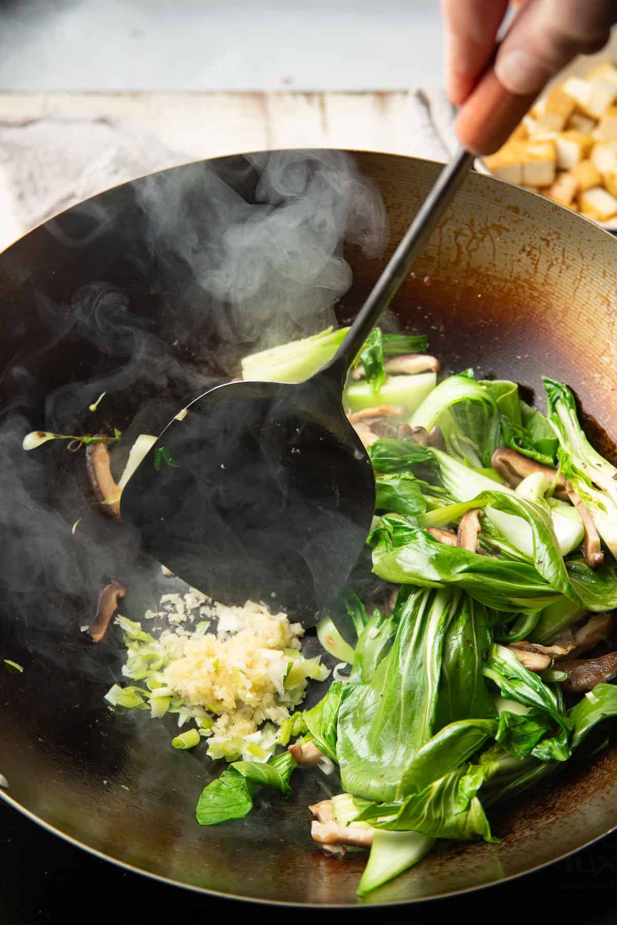 Hand with spatula sauteing garlic and ginger in a wok filled with vegetables.